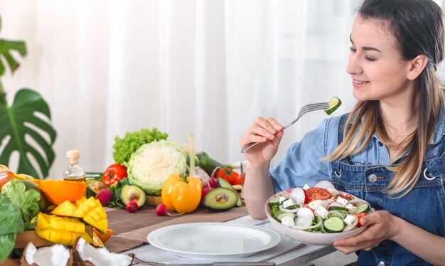 Mujer joven y feliz comiendo ensalada con verduras orgánicas en la mesa sobre un fondo claro, con ropa de mezclilla. El concepto de comida casera saludable.