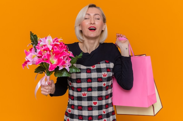 Mujer joven feliz y complacida en un hermoso vestido con ramo de flores y bolsas de papel con regalos celebrando el día internacional de la mujer de pie sobre la pared naranja