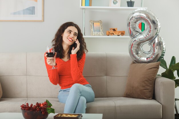 Mujer joven feliz y complacida en ropa casual sonriendo alegremente sentada en un sofá con una copa de vino hablando por teléfono móvil en la sala de estar luminosa celebrando el día internacional de la mujer el 8 de marzo