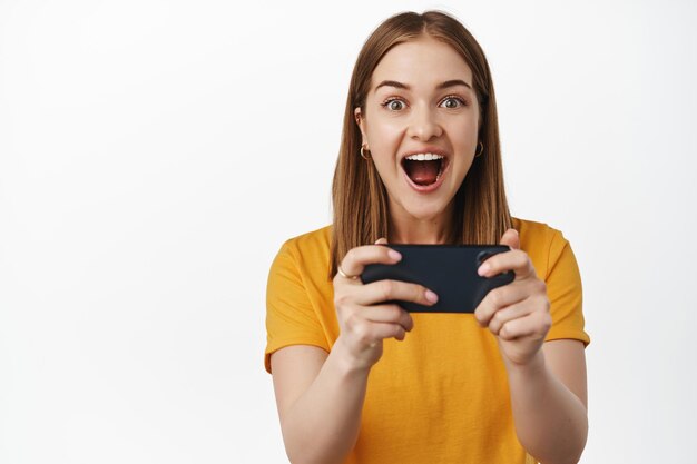 Mujer joven feliz y emocionada ganando en el teléfono móvil, sosteniendo el teléfono inteligente con ambas manos y gritando emocionada, viendo la transmisión de video en el teléfono celular, fondo blanco.