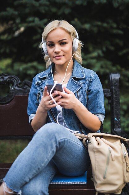 Mujer joven feliz escuchando música en auriculares y usando el teléfono inteligente mientras está sentado en el banco en la ciudad