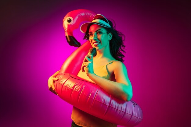 Mujer joven feliz en un flamenco de goma, gorra roja y gafas de sol con un helado en neón rosa de moda