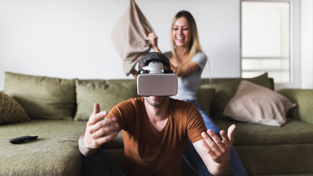 Mujer joven feliz golpeando a su novio con gafas de realidad virtual con cojín