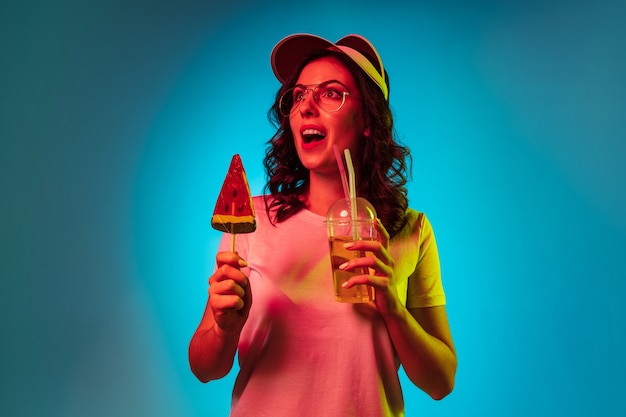 Mujer joven feliz en una gorra con dulces y bebida sobre estudio de neón azul de moda