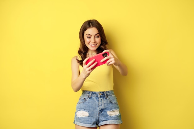 Mujer joven feliz jugando en el teléfono móvil, videojuego de teléfono inteligente, sonriendo y mirando la pantalla con entusiasmo, de pie sobre fondo amarillo