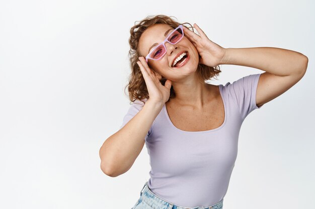 Mujer joven feliz y libre en gafas de sol, mirando con alegría, usando gafas y sonriendo, de pie en blanco