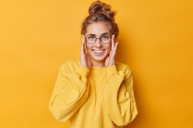 La mujer joven feliz mantiene las manos en la cara, las sonrisas muestran ampliamente los dientes blancos, usa anteojos redondos para ver un puente casual aislado sobre un fondo amarillo. Concepto de expresiones y emociones del rostro humano