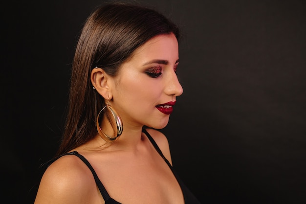 Mujer joven feliz con maquillaje brillante y joyas doradas en vestido negro posando