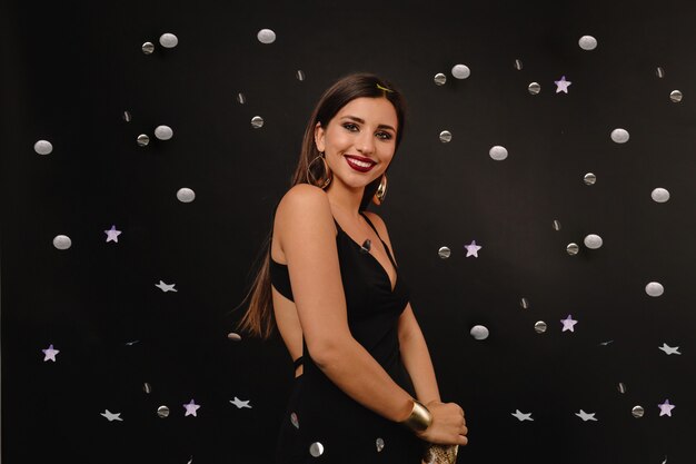 Mujer joven feliz con maquillaje brillante y joyas doradas en vestido negro posando