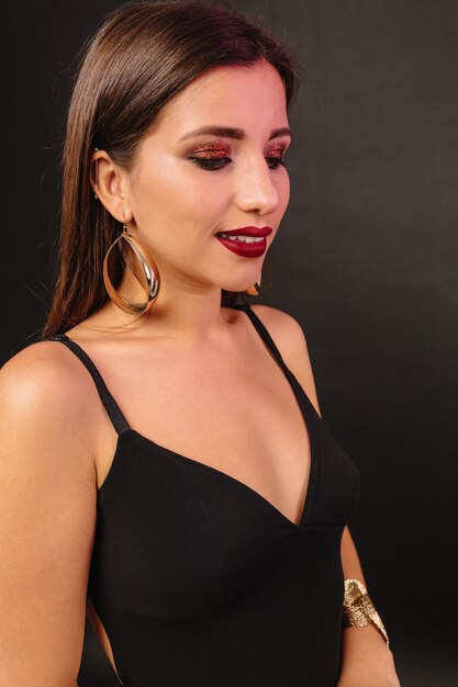 Mujer joven feliz con maquillaje brillante y joyas doradas en vestido negro posando