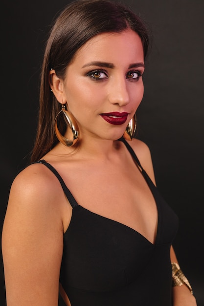 Mujer joven feliz con maquillaje brillante y joyas doradas en vestido negro posando