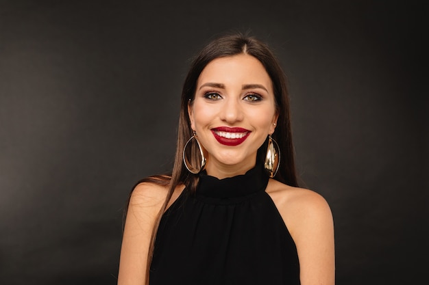 Mujer joven feliz con maquillaje brillante y joyas doradas en vestido negro posando