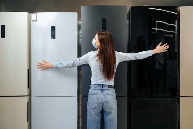 Mujer joven feliz en máscara apoyándose en su nuevo refrigerador en un centro comercial