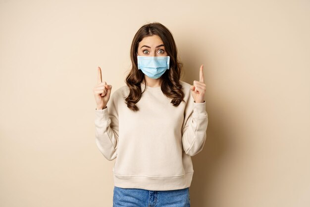 Mujer joven feliz con máscara médica de covid-19, señalando con el dedo hacia arriba, mostrando espacio de promoción o copia, de pie sobre fondo beige