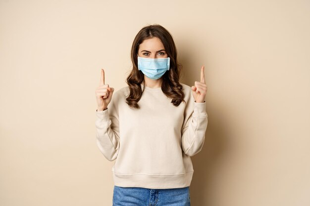 Mujer joven feliz con máscara médica de covid señalando con el dedo hacia arriba mostrando promoción o espacio de copia standin...