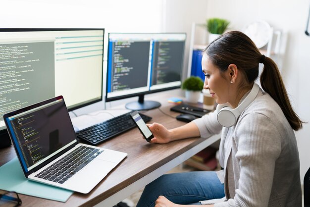Mujer joven feliz mirando su teléfono inteligente y enviando mensajes de texto mientras codifica en su computadora. Programador trabajando en el desarrollo de software en casa