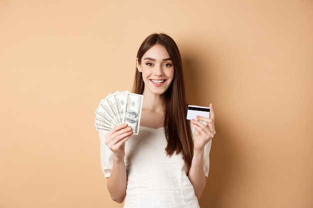 Mujer joven feliz mostrando billetes de un dólar y tarjeta de crédito de plástico sonriendo complacido haciendo dinero y compra ...