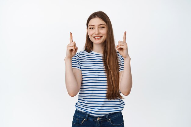 Mujer joven feliz mostrando información, señalando con el dedo hacia arriba y sonriendo complacido, demostrando pancarta de venta, de pie en blanco.