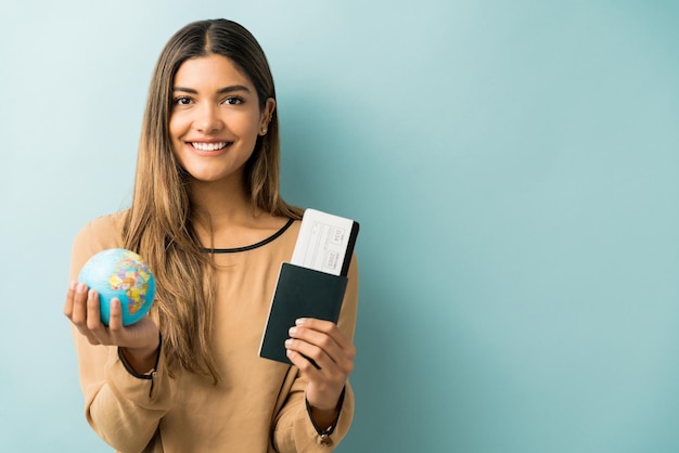 Mujer joven feliz con pasaporte y tarjeta de embarque con globo mientras está de pie contra el fondo azul