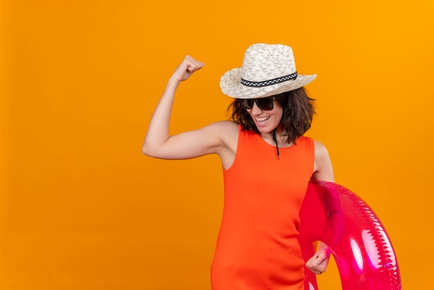 Foto gratuita una mujer joven feliz con el pelo corto en una camisa naranja con sombrero para el sol y gafas de sol sosteniendo un anillo inflable mostrando gesto de fuerza