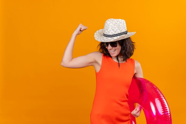 Una mujer joven feliz con el pelo corto en una camisa naranja con sombrero para el sol y gafas de sol sosteniendo un anillo inflable mostrando gesto de fuerza