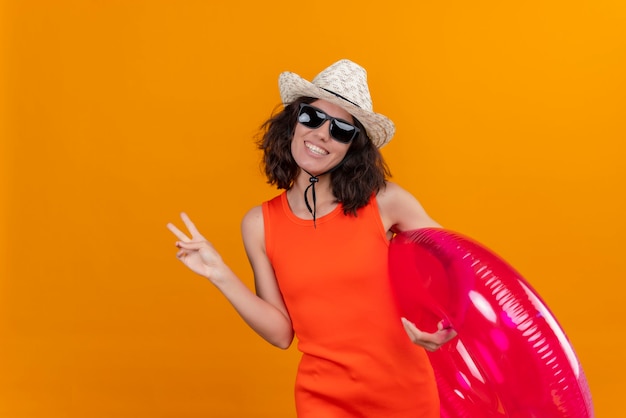 Una mujer joven feliz con el pelo corto en una camisa naranja con sombrero para el sol y gafas de sol sosteniendo un anillo inflable que muestra el gesto de dos dedos