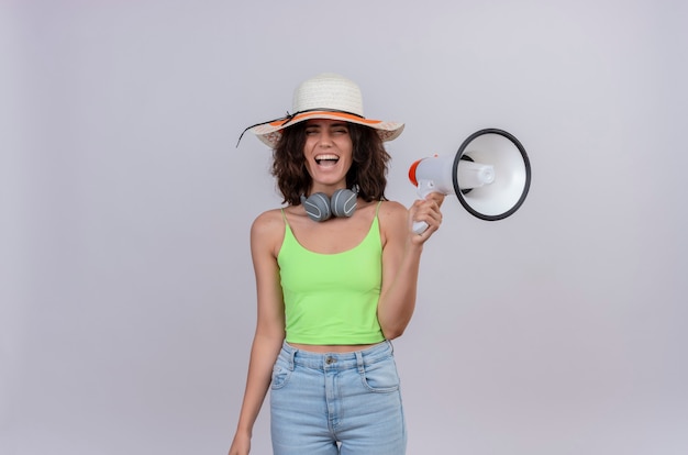 Una mujer joven feliz con el pelo corto en la parte superior de la cosecha verde en auriculares con sombrero para el sol sonriendo y sosteniendo el megáfono sobre un fondo blanco.