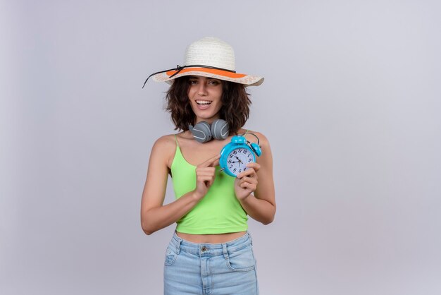 Una mujer joven feliz con el pelo corto en la parte superior de la cosecha verde con sombrero para el sol sosteniendo el despertador azul sobre un fondo blanco.