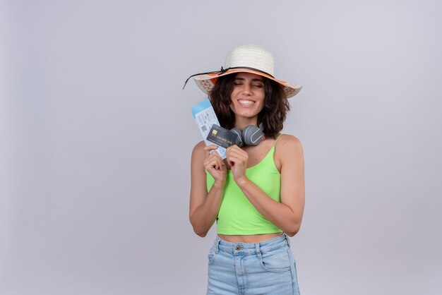 Una mujer joven feliz con el pelo corto en la parte superior verde con sombrero para el sol con billetes de avión y tarjeta de crédito sobre un fondo blanco