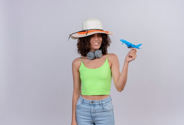 Una mujer joven feliz con el pelo corto en verde crop top con sombrero para el sol sosteniendo avión de juguete azul sobre un fondo blanco.