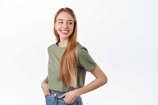 Mujer joven feliz con el pelo largo y rubio, gira la cabeza detrás del hombro y sonriendo al logotipo del anuncio a un lado, de pie sobre una pared blanca