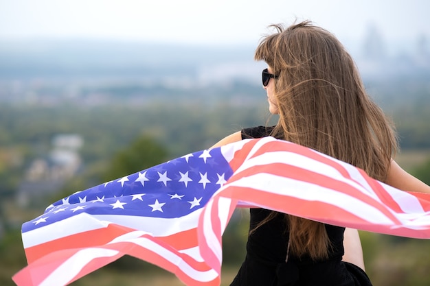 Mujer joven feliz con el pelo largo sosteniendo ondeando en el viento la bandera nacional americana en sus hombros descansando al aire libre disfrutando de un cálido día de verano.