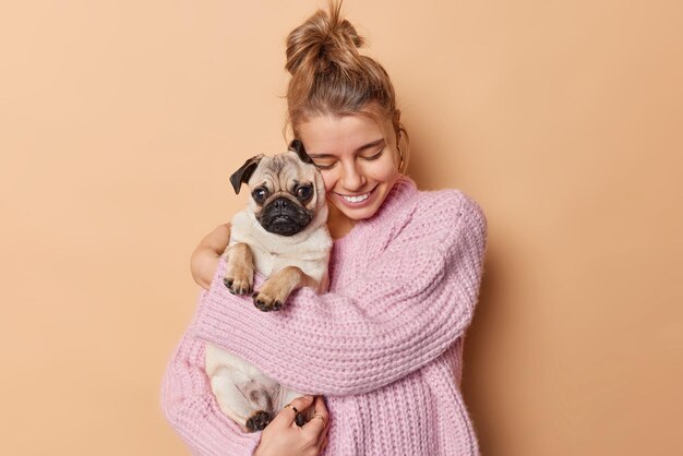 Una mujer joven feliz con el pelo peinado abraza a un perro pug que se divierte siendo mamá de un cachorro que viste un suéter de punto aislado sobre un fondo beige Amante de las mascotas con animales domésticos