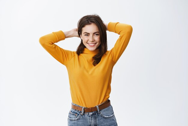 Mujer joven feliz de pie relajada con las manos detrás de la cabeza con tiempo libre descansando y sonriendo de pie sobre fondo blanco.