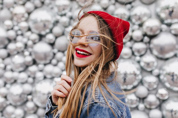 Mujer joven feliz posando delante de bolas de discoteca y riendo. Retrato de niña caucásica romántica en moda sombrero rojo y gafas de sol.