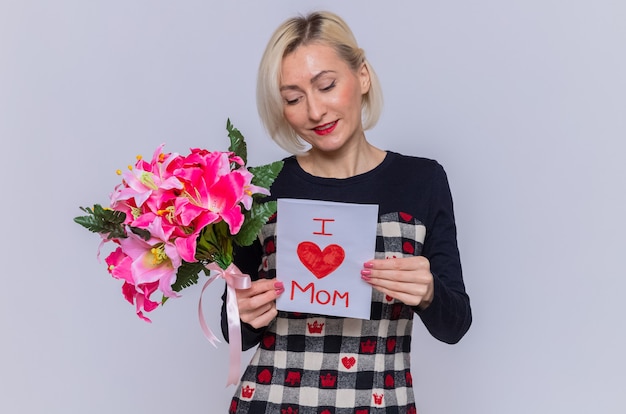 Mujer joven feliz y positiva en un hermoso vestido con tarjeta de felicitación y ramo de flores sonriendo alegremente celebrando el día internacional de la mujer de pie sobre una pared blanca