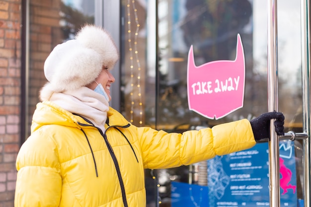 Mujer joven feliz en la puerta del restaurante en un día frío de invierno, rotulación, comida para llevar.