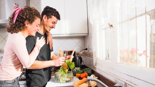 Mujer joven feliz que abraza a su marido de detrás que prepara la ensalada en el cuenco