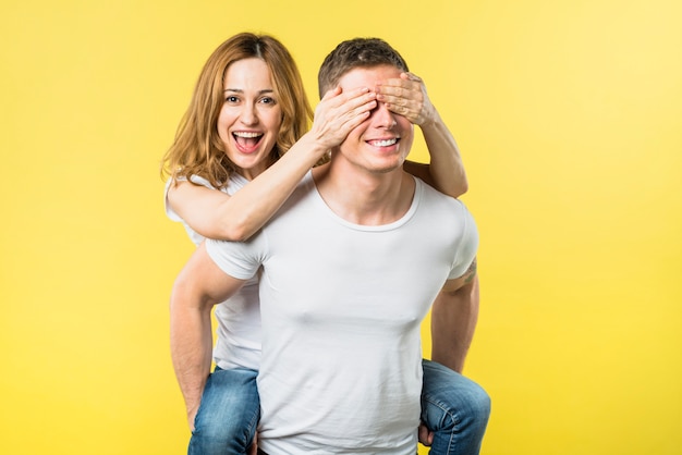 Mujer joven feliz que cubre los ojos mientras monta la espalda de su novio contra el fondo amarillo