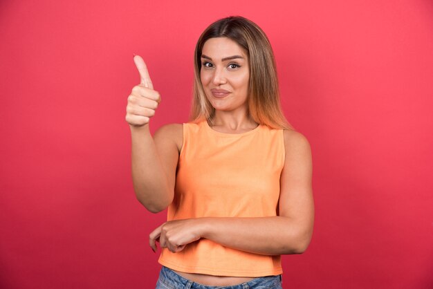 Mujer joven feliz que hace los pulgares para arriba en la pared roja.