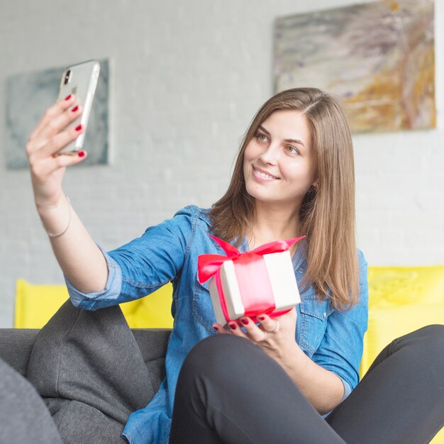 Mujer joven feliz que lleva a cabo el presente de cumpleaños que toma el selfie con el teléfono móvil