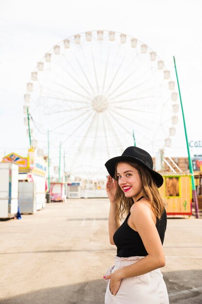 Mujer joven feliz que mira sobre hombro con su mano en bolsillo el parque de atracciones