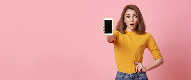 Mujer joven feliz que muestra en el éxito del gesto de la mano y del teléfono móvil de la pantalla en blanco aislado sobre fondo rosado.