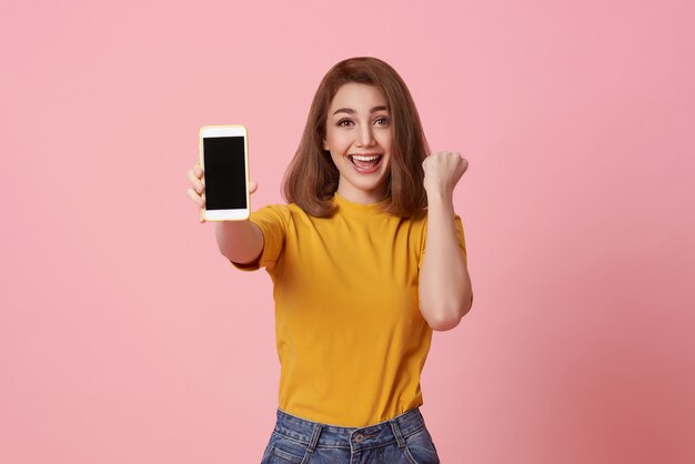 Mujer joven feliz que muestra en el éxito del gesto de la mano y del teléfono móvil de la pantalla en blanco aislado sobre fondo rosado.