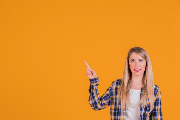 Mujer joven feliz que señala su dedo hacia arriba contra un fondo naranja