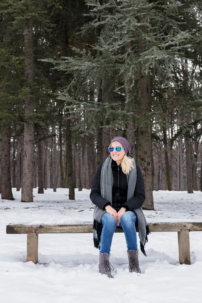 Mujer joven feliz que se sienta en el banco de madera que lleva la ropa caliente en invierno