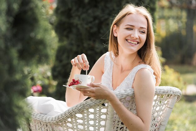 Mujer joven feliz que se sienta en la silla blanca que revuelve el café con la cuchara