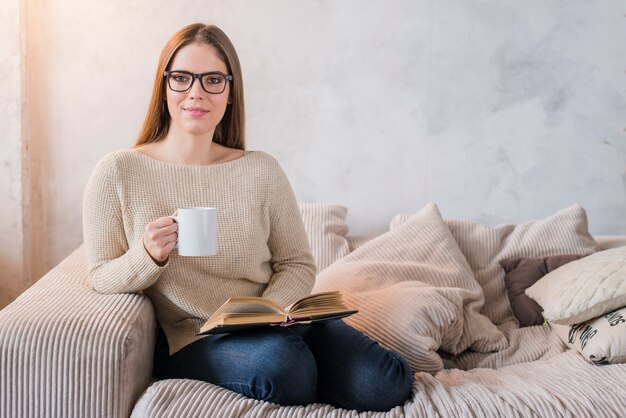 Mujer joven feliz que se sienta en el sofá que sostiene el libro y la taza de café en manos