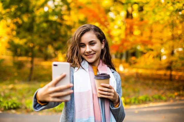 Mujer joven feliz que sostiene el café para llevar, tomando un selfie en el teléfono inteligente, al aire libre en otoño.