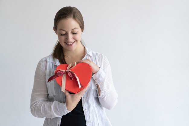 Mujer joven feliz que sostiene la caja de regalo en forma de corazón roja